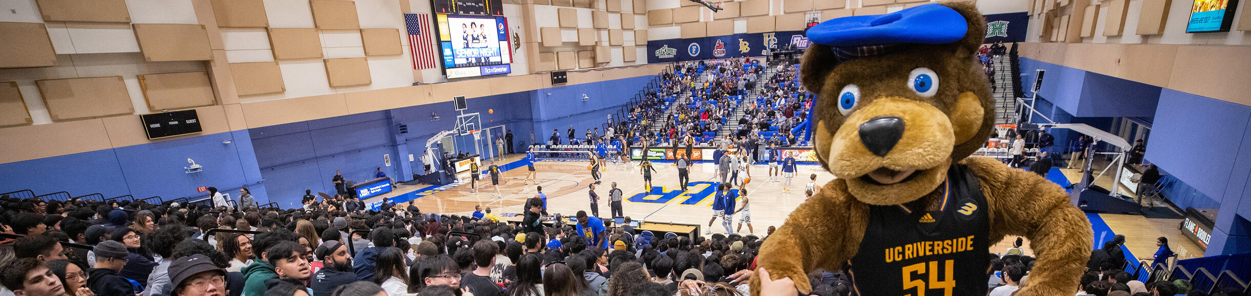 Los estudiantes de UC Riverside ven un partido de baloncesto en casa mientras la mascota de UCR Scotty entretiene a la multitud